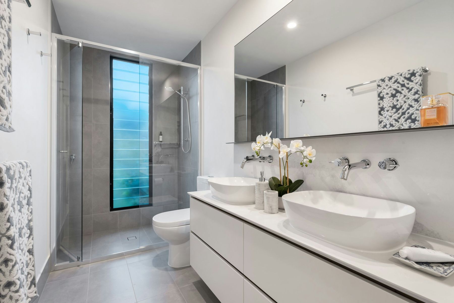 Bright white double vanity bathroom with glass shower and orchids — The Magnolia Bath, Keller